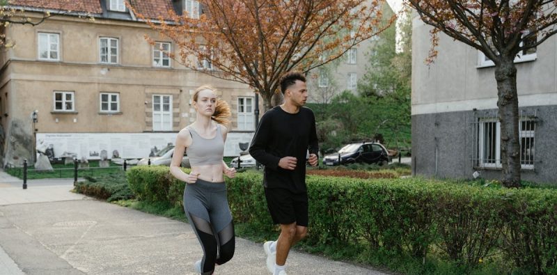 Two adults jogging on a sidewalk in an urban setting surrounded by spring foliage.