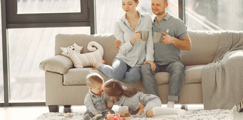 Cheerful family smiling while spending weekend together sitting parents sitting on comfortable sofa while children resting on carpet and playing
